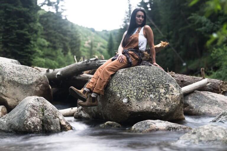 A hiker sitting on a large rock by a stream with their Ridgeway Chelsea boots