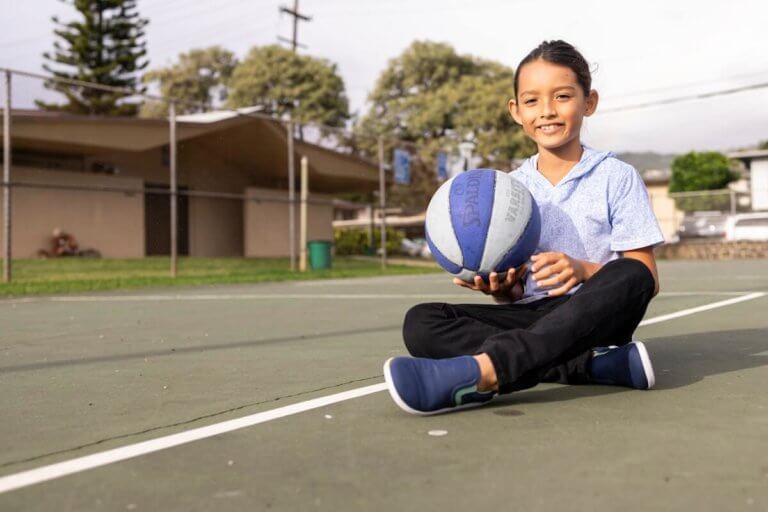 A boy sitting on a basketball court in his Dillon Canvas Slip-On Youth shoes.